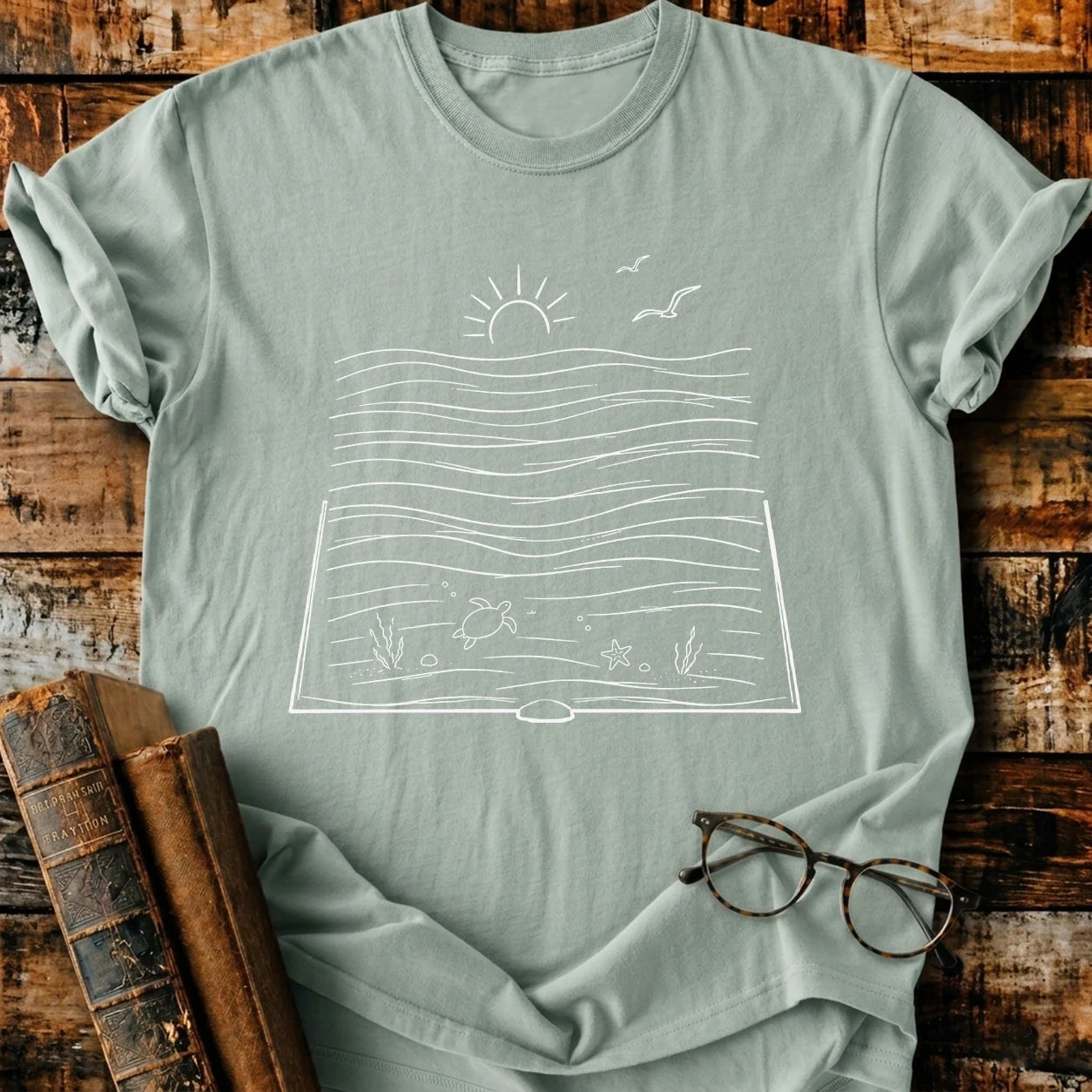 Sage green Comfort Colors t-shirt with a white "Bookish Ocean" graphic, featuring a sun, waves, turtle, and starfish.