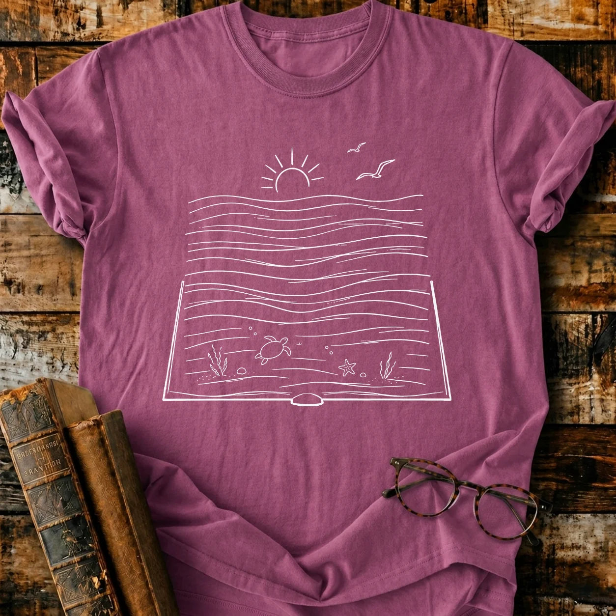 Bookish Ocean T-shirt: Garment-dyed berry Comfort Colors tee with white book-ocean graphic, turtle, sun, and gulls.