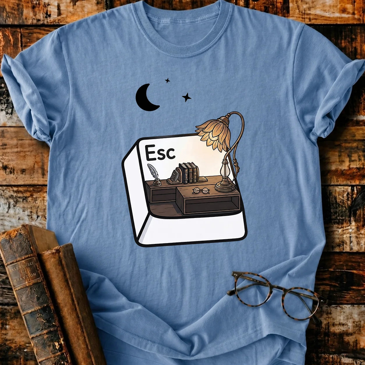 Light blue Comfort Colors Escape T-shirt with a desk, books, lamp, and moon graphic.