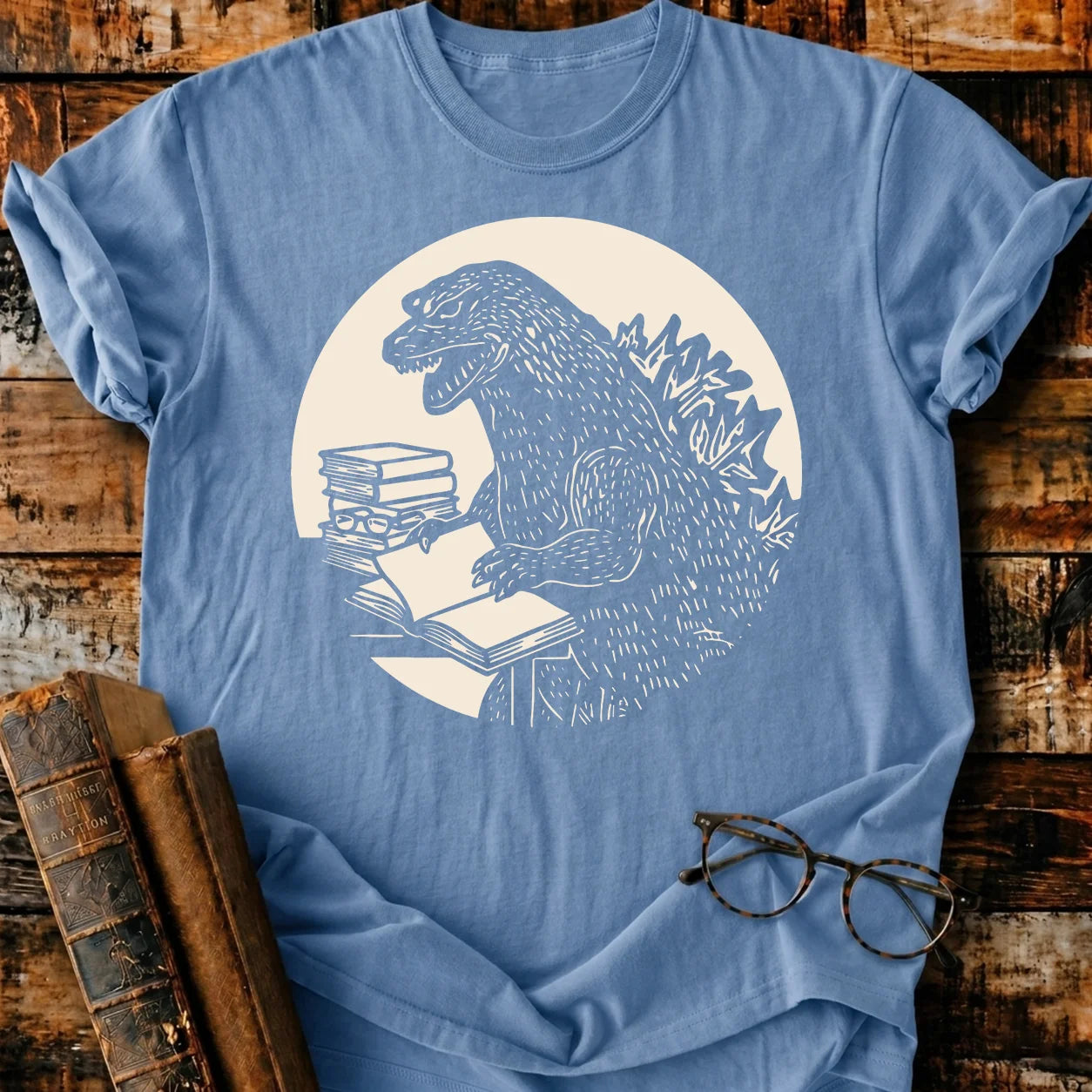Light blue Comfort Colors 1717 t-shirt with a white graphic of Godzilla reading a book.