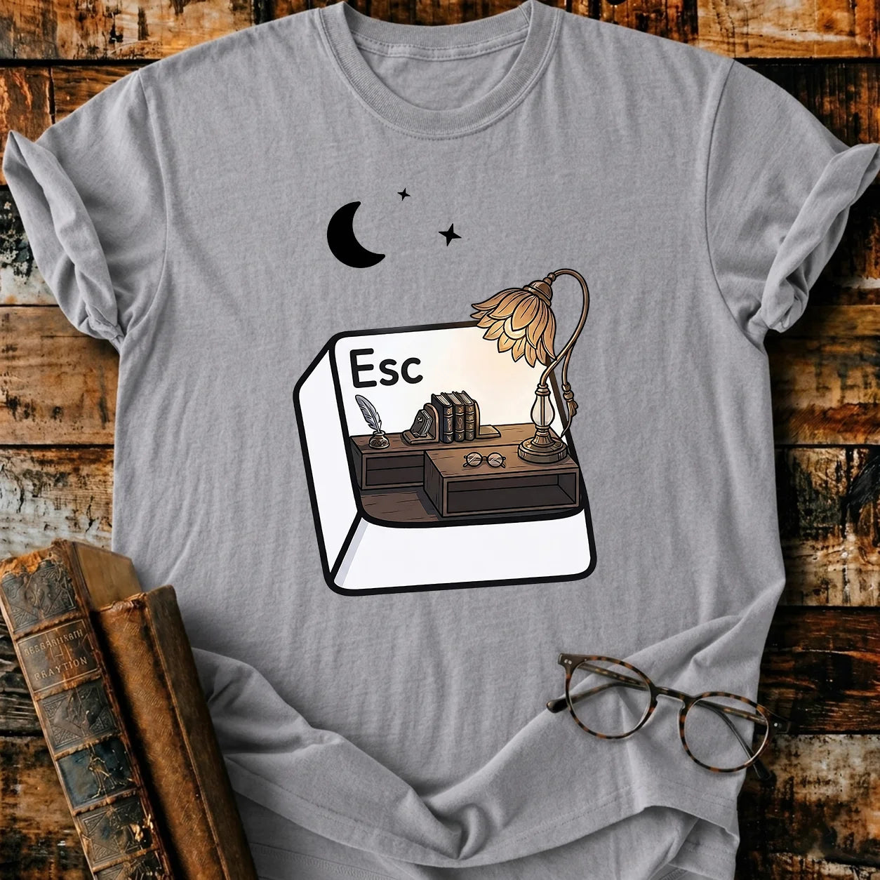 Gray Comfort Colors Escape T-shirt with a desk, lamp, books, and moon design on a wooden background.